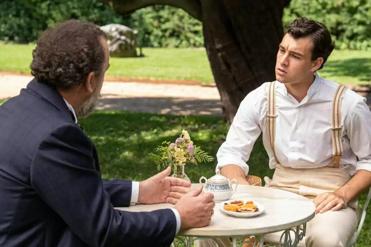 Curro y Alonso conversan en una mesa al aire libre en un jardín, uno de ellos lleva tirantes y una camisa blanca mientras el otro viste un traje oscuro, en la serie La Promesa. Curro y Alonso conversan en una mesa al aire libre en un jardín, uno de ellos lleva tirantes y una camisa blanca mientras el otro viste un traje oscuro, en la serie La Promesa.