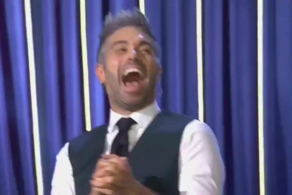 Dani Martínez riendo con entusiasmo frente a un fondo de cortinas azules en El Gran Show. Dani Martínez riendo con entusiasmo frente a un fondo de cortinas azules en El Gran Show.