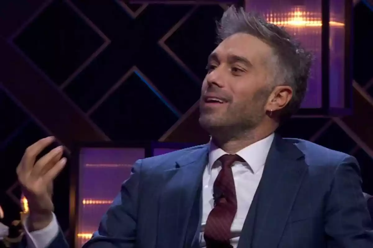 Dani Martínez con traje y corbata sonríe mientras gesticula con la mano en un ambiente iluminado en el plató de Martínez y Hermanos. Dani Martínez con traje y corbata sonríe mientras gesticula con la mano en un ambiente iluminado en el plató de Martínez y Hermanos.