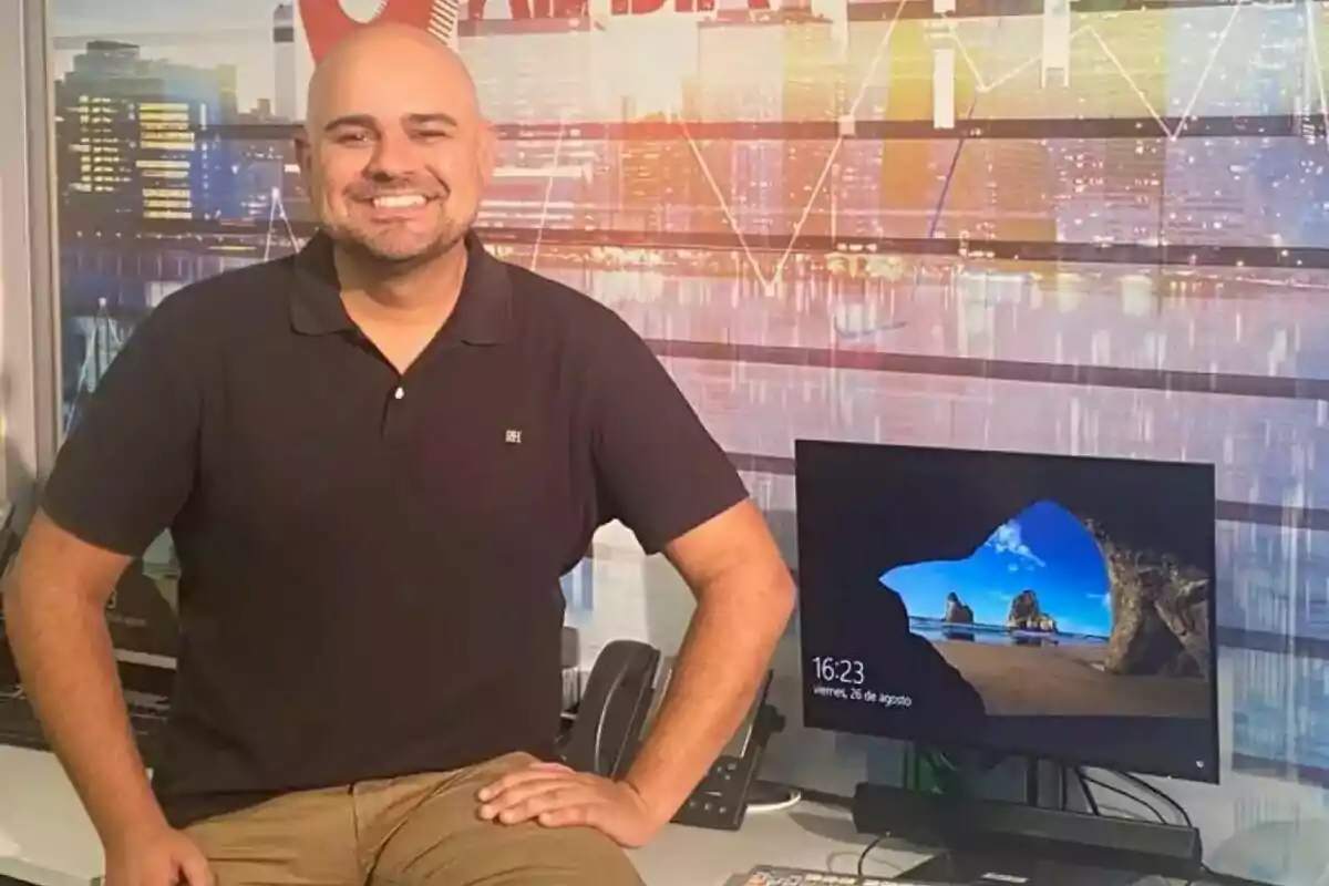 Daniel Fernández sonriente con camisa oscura está sentado en un escritorio junto a una computadora con una imagen de fondo de playa en la pantalla. Daniel Fernández sonriente con camisa oscura está sentado en un escritorio junto a una computadora con una imagen de fondo de playa en la pantalla.