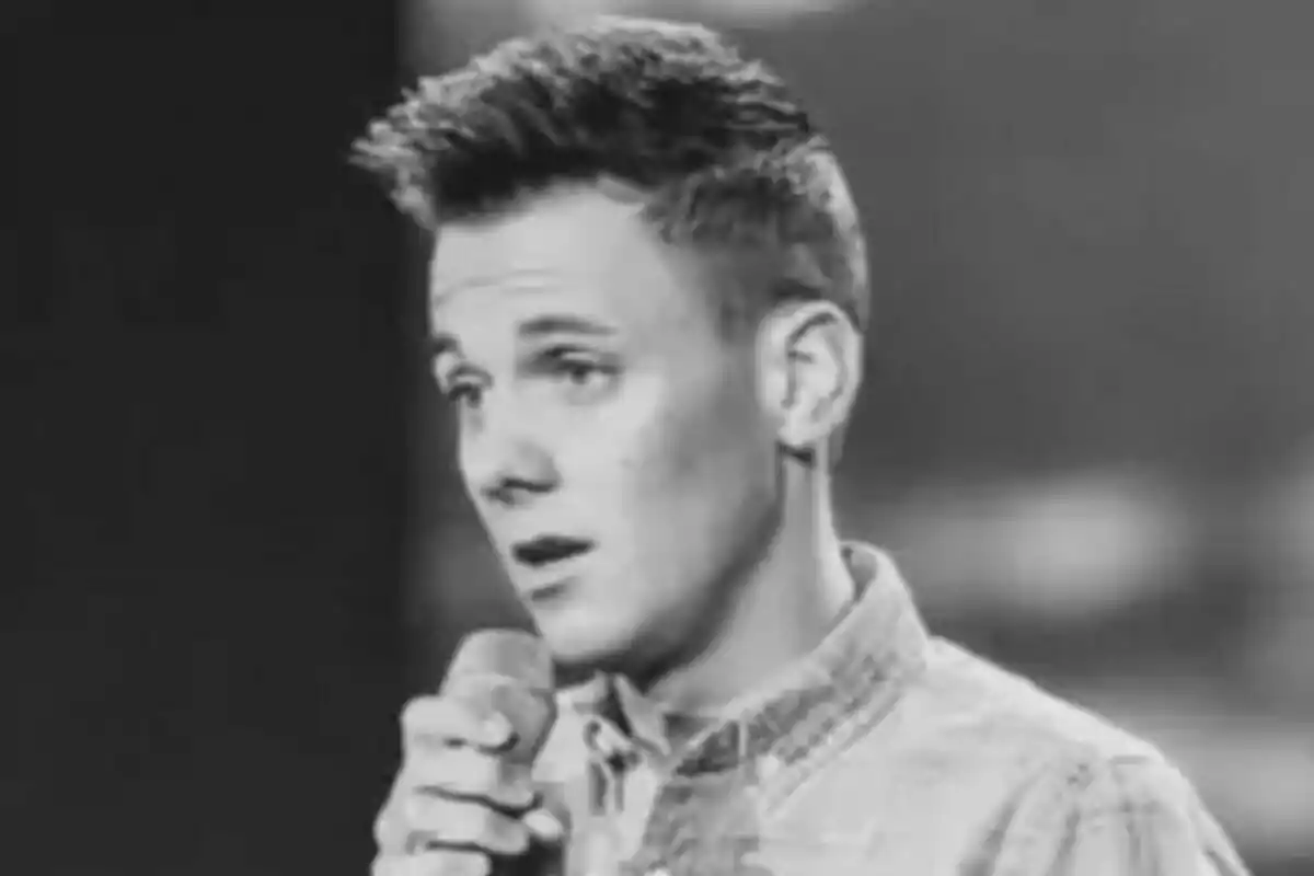 Danny Dearden con camisa de cuadros sostiene un micrófono mientras parece estar cantando o hablando en un escenario.