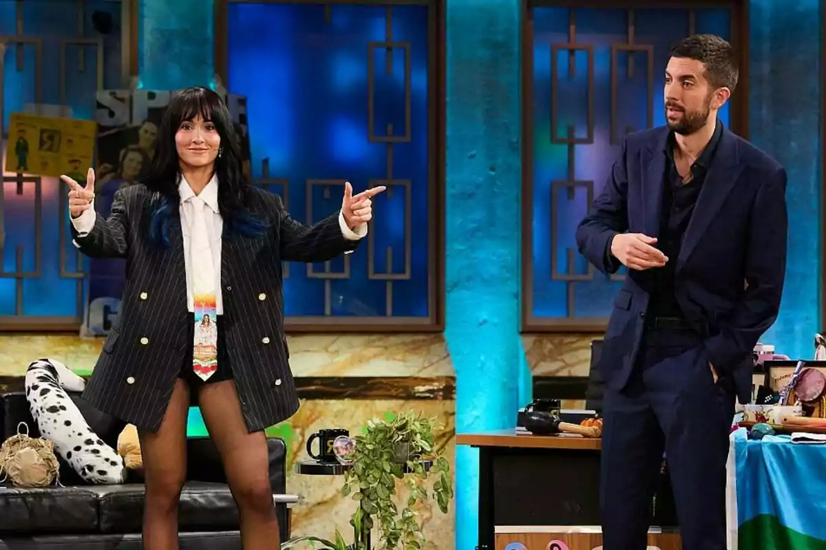 Aitana Ocaña y David Broncano están en La Revuelta, ella lleva un traje con chaqueta a rayas y corbata colorida, mientras que él viste un traje oscuro; el fondo tiene luces azules y decoraciones. Aitana Ocaña y David Broncano están en La Revuelta, ella lleva un traje con chaqueta a rayas y corbata colorida, mientras que él viste un traje oscuro; el fondo tiene luces azules y decoraciones.