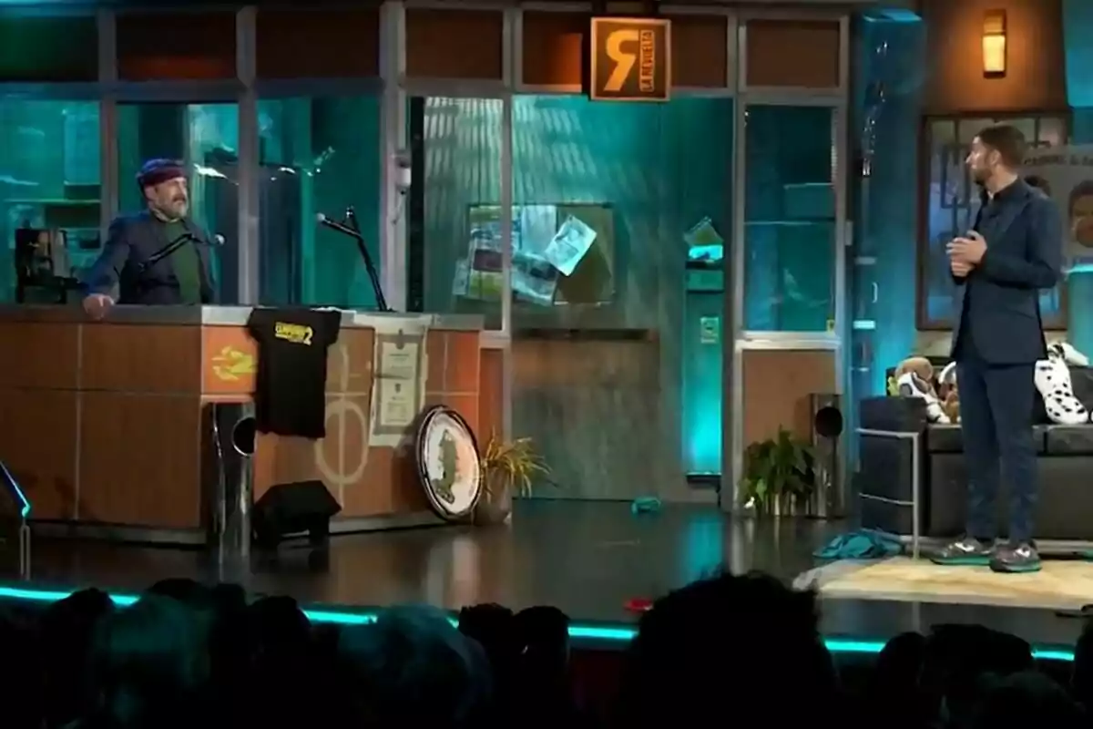 Ricardo Castella de pie detrás de un escritorio con micrófonos y una camiseta colgada en La Revuelta, mientras David Broncano está de pie a la derecha en un escenario con decoración de oficina y público al frente. Ricardo Castella de pie detrás de un escritorio con micrófonos y una camiseta colgada en La Revuelta, mientras David Broncano está de pie a la derecha en un escenario con decoración de oficina y público al frente.