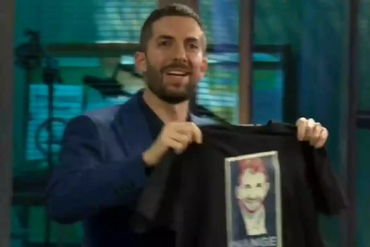 David Broncano con barba y chaqueta azul en La Revuelta sostiene una camiseta negra con la cara de Pablo Motos impresa en el frente. David Broncano con barba y chaqueta azul en La Revuelta sostiene una camiseta negra con la cara de Pablo Motos impresa en el frente.