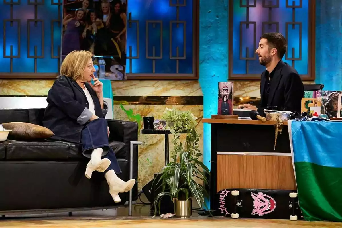 Carmen Machi sentada en un sofá negro conversa con David Broncano detrás de un escritorio en La Revuelta decorado con plantas y un monopatín. Carmen Machi sentada en un sofá negro conversa con David Broncano detrás de un escritorio en La Revuelta decorado con plantas y un monopatín.