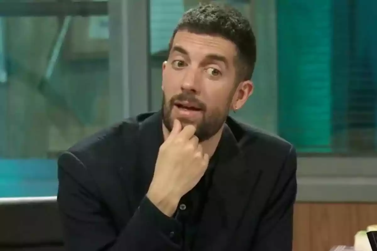 David Broncano con barba y cabello corto está sentado, apoyando su mano en el mentón, mientras parece estar hablando o pensando en un entorno interior en 'La Revuelta'.