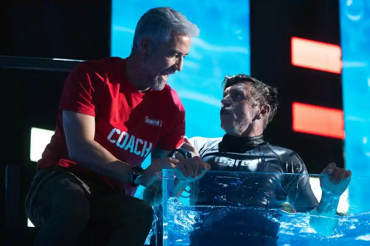 Manuel Díaz El Cordobés y su instructor de El Desafío interactúan en un entorno acuático, una de ellas está parcialmente sumergida en agua mientras la otra, con una camiseta roja que dice Manuel Díaz El Cordobés y su instructor de El Desafío interactúan en un entorno acuático, una de ellas está parcialmente sumergida en agua mientras la otra, con una camiseta roja que dice