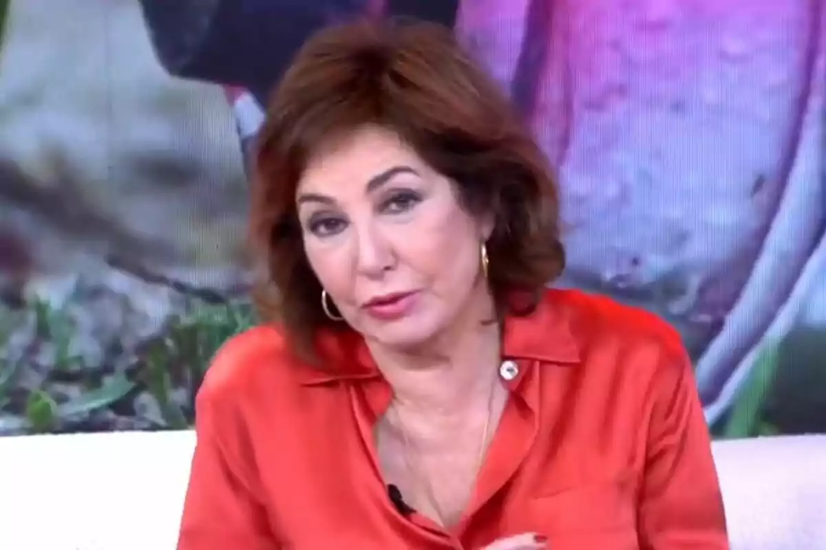 Ana Rosa Quintana con cabello castaño y una blusa naranja, sentada frente a un fondo colorido en el plató de TardeAR.