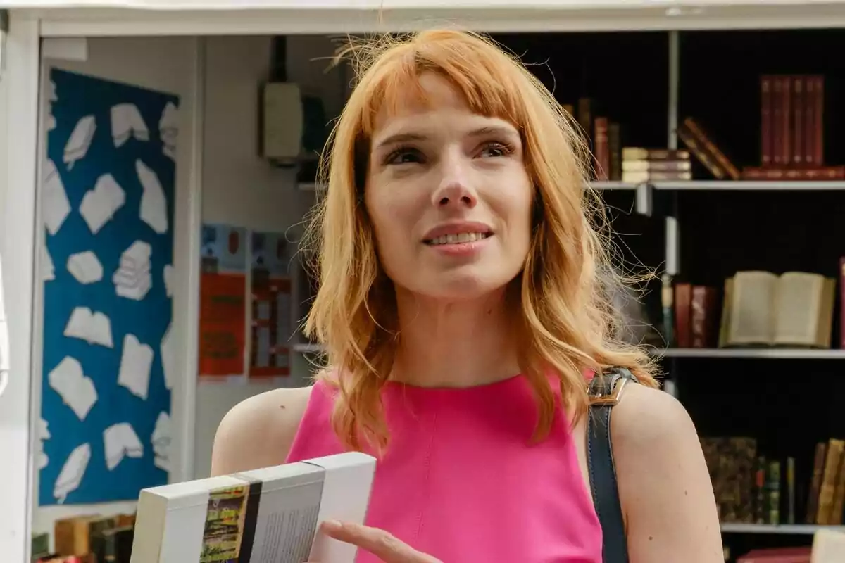 Diana Gómez con vestido rosa sosteniendo un libro frente a una librería en la serie Valeria.