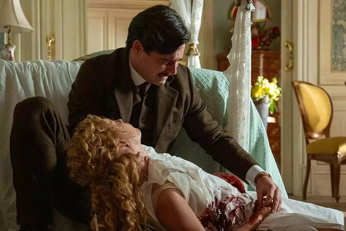 Arturo Sancho con bigote sostiene a Ana Garcés herida en una habitación elegantemente decorada en La Promesa. Arturo Sancho con bigote sostiene a Ana Garcés herida en una habitación elegantemente decorada en La Promesa.