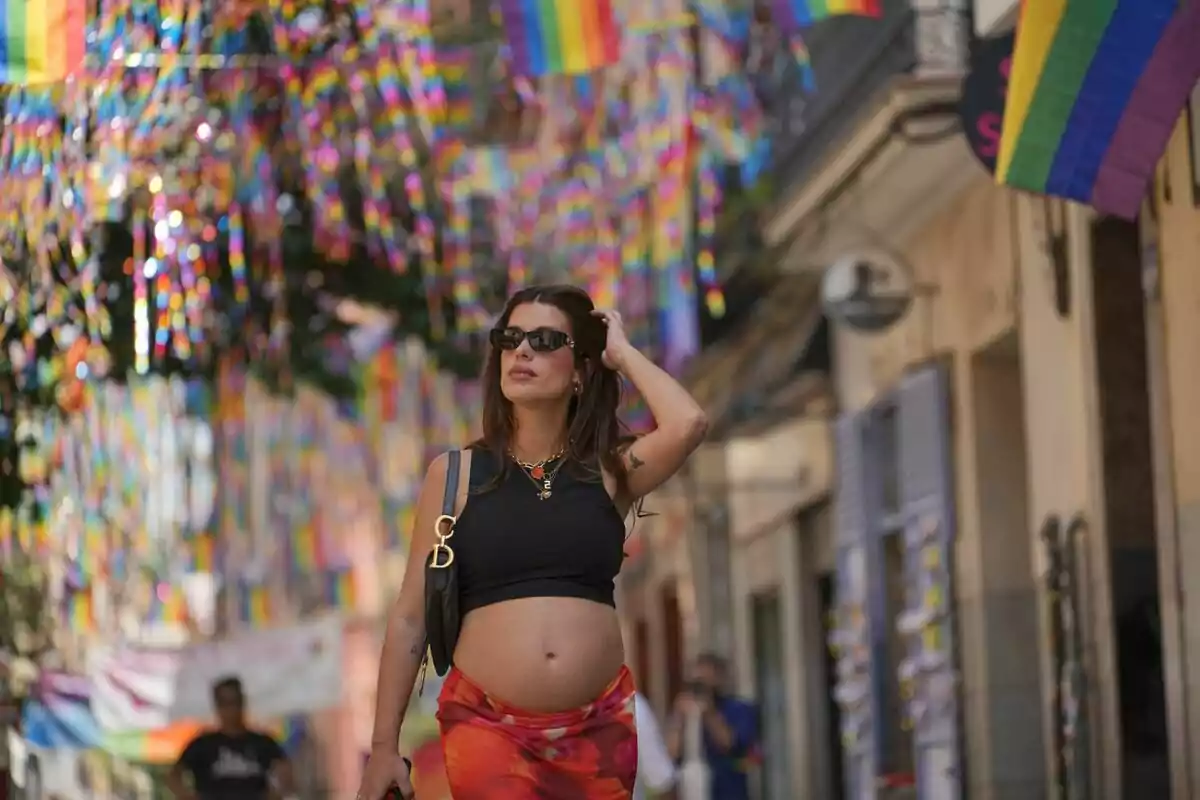 Dulceida embarazada camina por una calle decorada con banderas de arcoíris, luciendo gafas de sol y ropa veraniega en Dulceida al Desnudo.