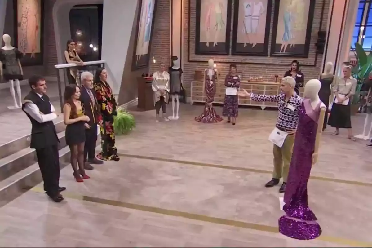 Los jueces está en un estudio de diseño de moda, observando un maniquí con un vestido brillante de color púrpura, mientras que otras personas están de pie cerca de maniquíes y bocetos de moda en las paredes. Los jueces está en un estudio de diseño de moda, observando un maniquí con un vestido brillante de color púrpura, mientras que otras personas están de pie cerca de maniquíes y bocetos de moda en las paredes.