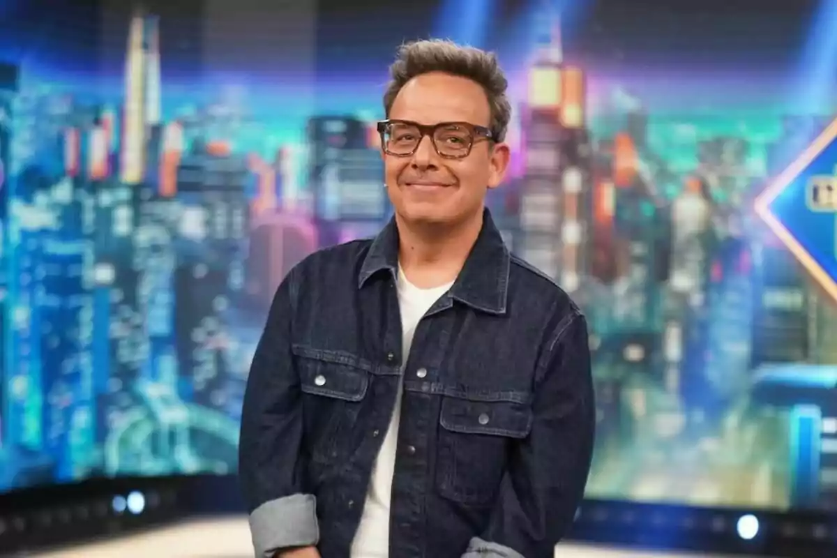 Àngel Llàser amb ulleres i jaqueta texana somriu davant d'un fons urbà il·luminat a 'El Hormiguero'.