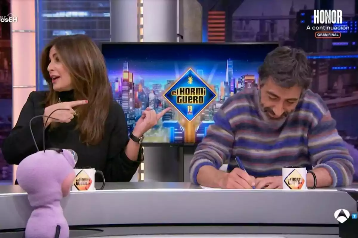 En un estudio de televisión, Nuria Roca gesticula mientras Juan del Val escribe, con un muñeco morado en primer plano y un logotipo de En un estudio de televisión, Nuria Roca gesticula mientras Juan del Val escribe, con un muñeco morado en primer plano y un logotipo de