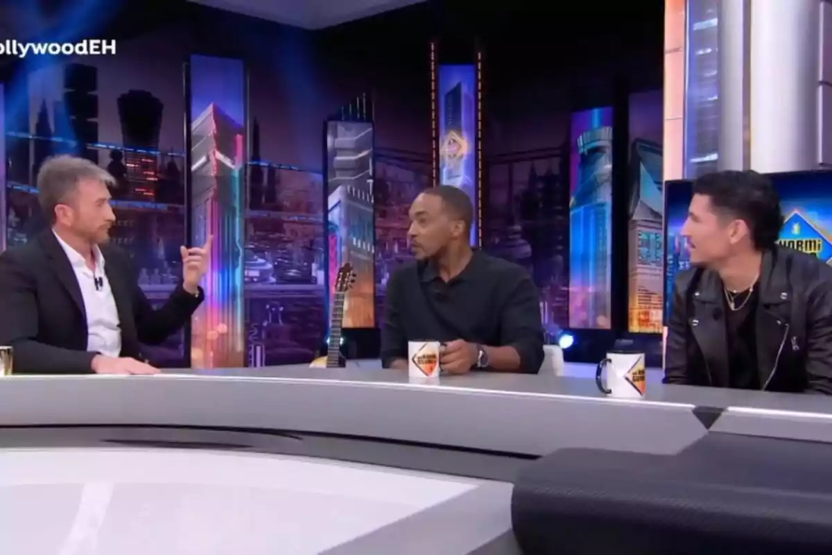 Pablo Motos, Anthony Mackie y Danny Ramírez sentados en 'El Hormiguero', conversando alrededor de una mesa con tazas de café, con un fondo de luces y edificios iluminados.