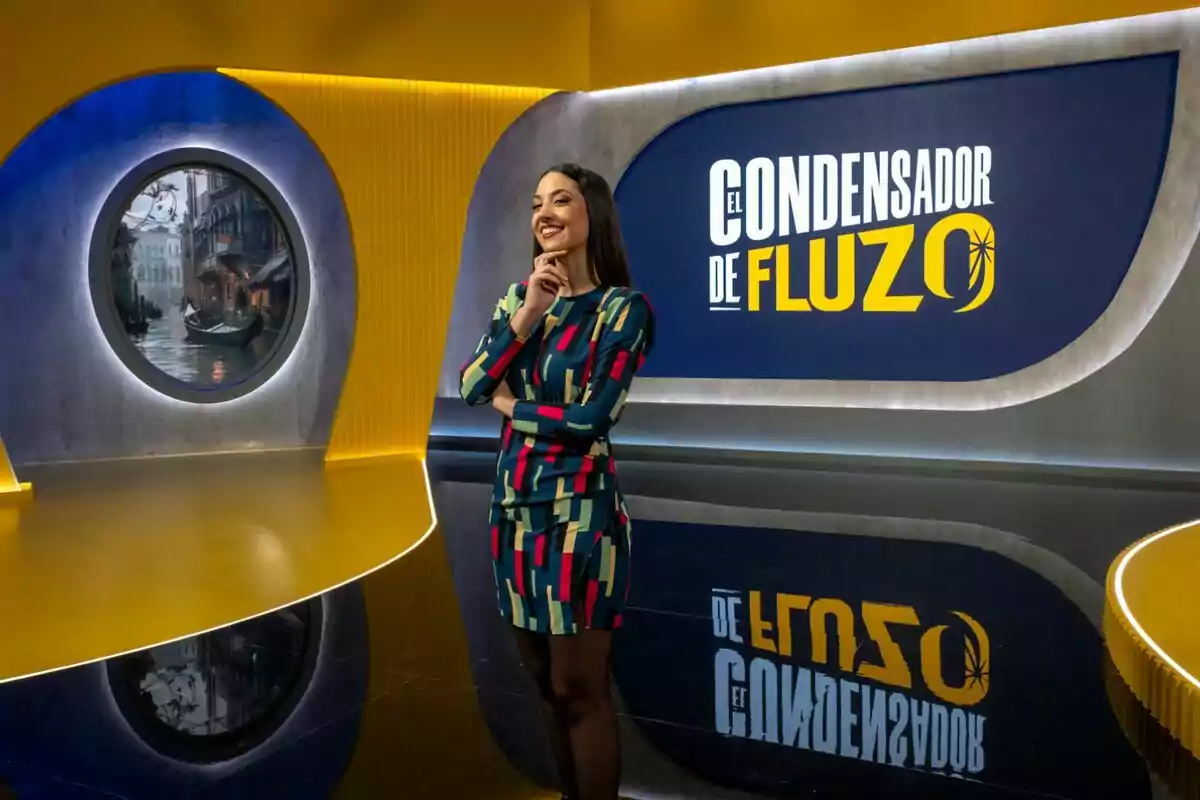 Maya Pixelskaya sonriente en un set de televisión con un diseño moderno y colorido, con un letrero que dice Maya Pixelskaya sonriente en un set de televisión con un diseño moderno y colorido, con un letrero que dice