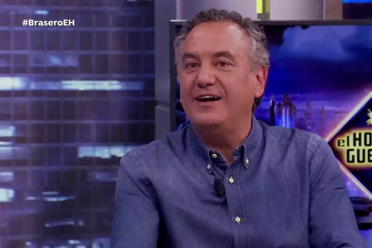 Roberto Brasero con camisa de mezclilla sonríe mientras está sentado en el set de televisión con el logo de Roberto Brasero con camisa de mezclilla sonríe mientras está sentado en el set de televisión con el logo de