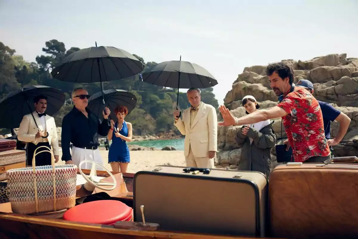 Un grupo de personas encabezado por José Coronado con sombrillas negras está de pie en una playa junto a un bote, mientras un hombre con camisa hawaiana parece estar dando instrucciones en el rodaje de la serie El Problema Final.