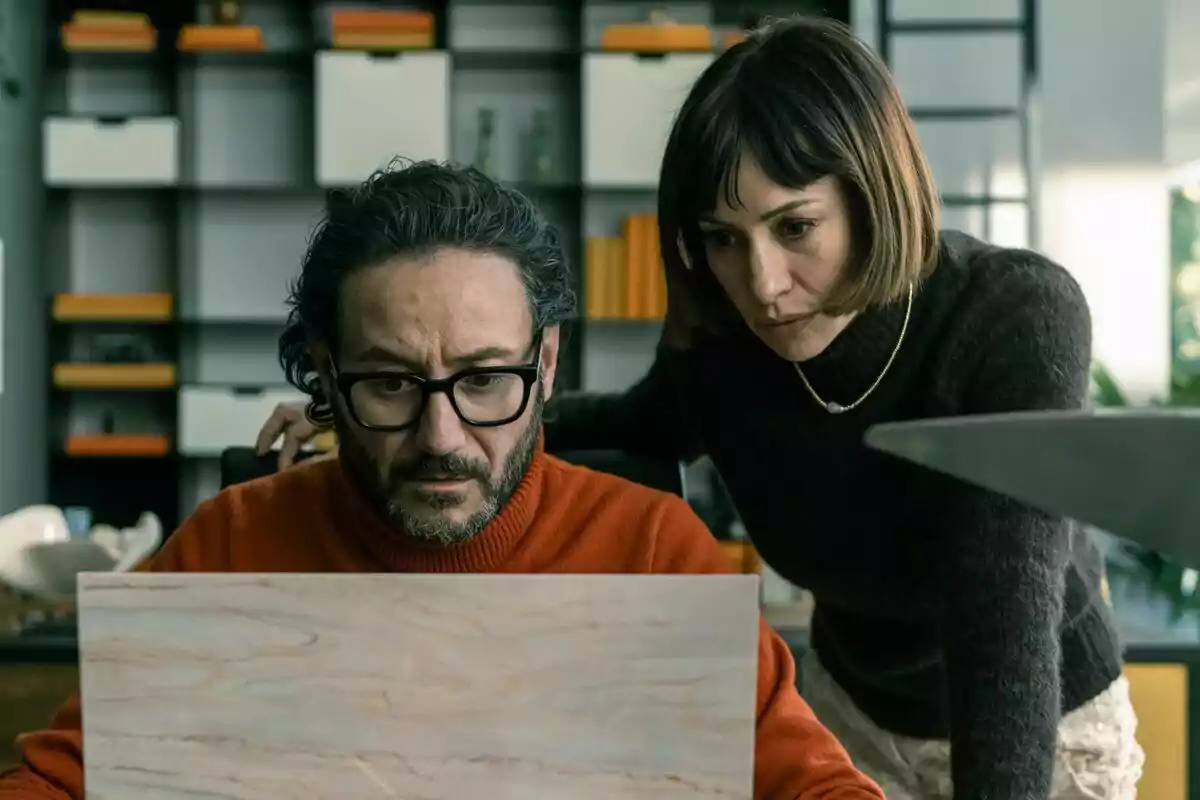 Carlos Santos con gafas y suéter naranja mira preocupado la pantalla de una laptop mientras una mujer de cabello corto y suéter oscuro lo observa atentamente desde su lado en una oficina moderna en El Refugio Atómico.