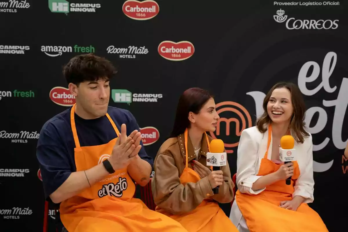 Andrea Garte, Viruzz y Paula Feel con delantales naranjas participan en un evento de cocina, sosteniendo micrófonos y sentados frente a un fondo con logotipos de marcas. Andrea Garte, Viruzz y Paula Feel con delantales naranjas participan en un evento de cocina, sosteniendo micrófonos y sentados frente a un fondo con logotipos de marcas.