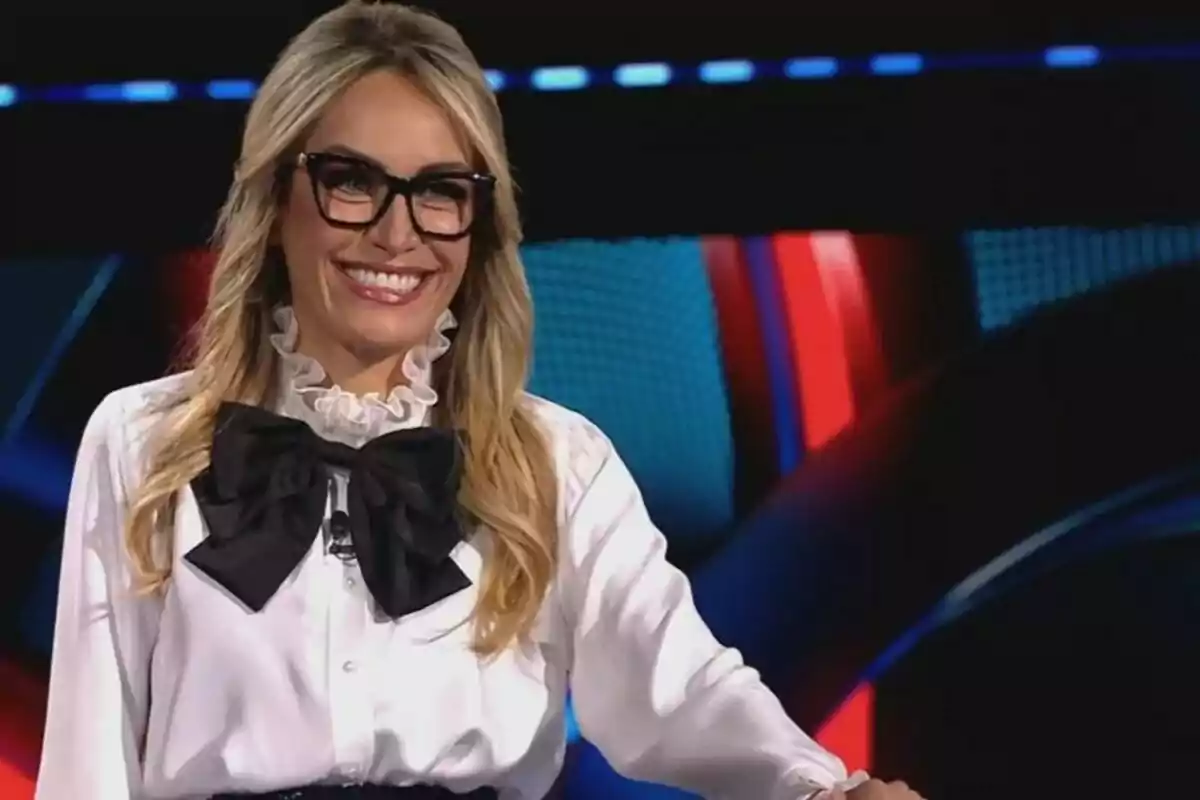 Luján Argüelles rubia con gafas y blusa blanca con un gran lazo negro, sonriendo frente a un fondo colorido en el plató de El Rival Más Débil.