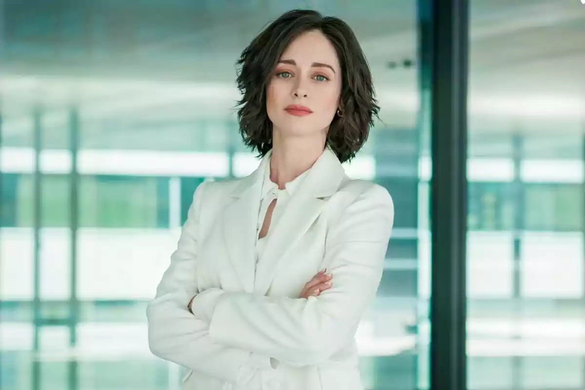 Elena Rivera amb cabell curt i fosc vestida amb un vestit blanc, dempeus amb els braços creuats en un entorn modern i lluminós a la sèrie Perdent el Judici.