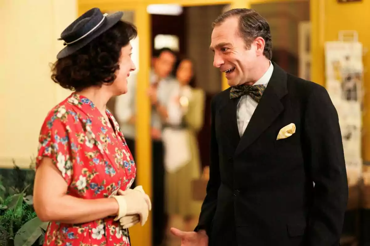 Elías i Leonora conversant alegrement en un ambient interior, una dona amb un vestit floral i un barret i un home amb vestit i corbata a la sèrie La Moderna. Elías i Leonora conversant alegrement en un ambient interior, una dona amb un vestit floral i un barret i un home amb vestit i corbata a la sèrie La Moderna.