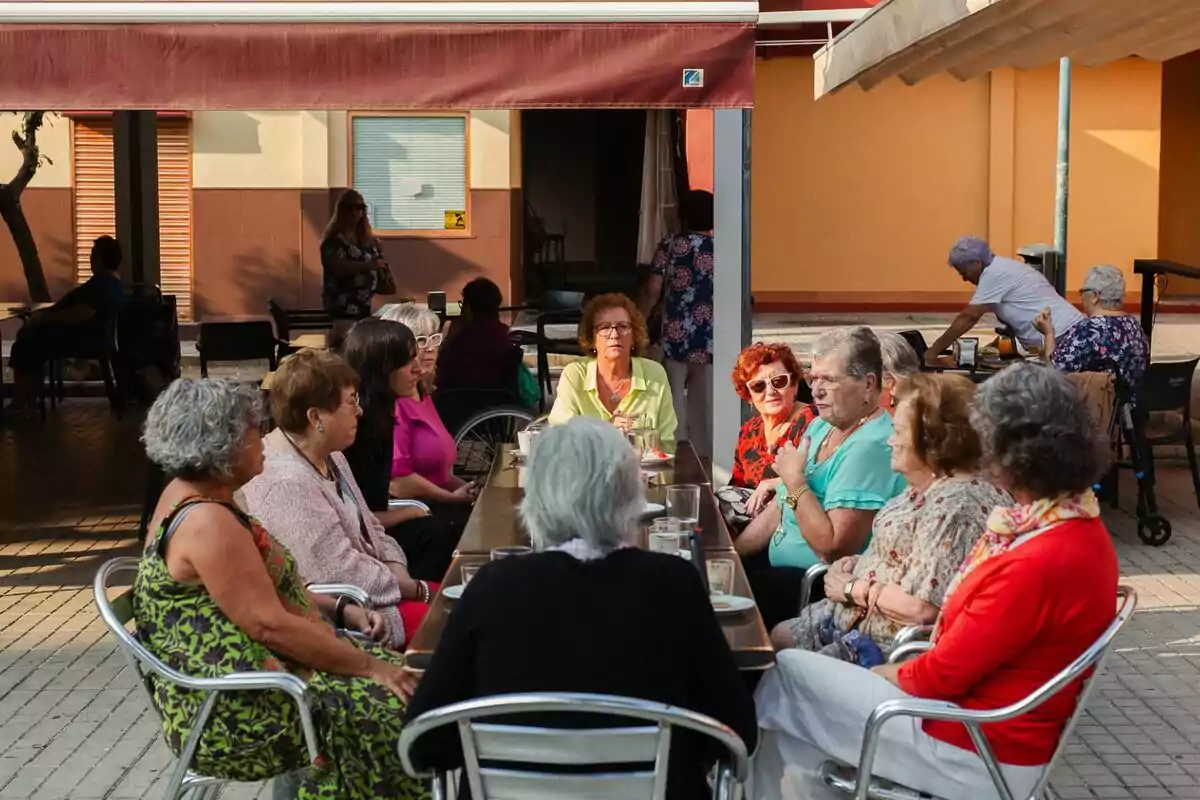 Un grupo de personas mayores se encuentra sentado alrededor de una mesa al aire libre en una terraza, conversando y disfrutando de una reunión social en un día soleado en el documental Ellas en la ciudad. Un grupo de personas mayores se encuentra sentado alrededor de una mesa al aire libre en una terraza, conversando y disfrutando de una reunión social en un día soleado en el documental Ellas en la ciudad.