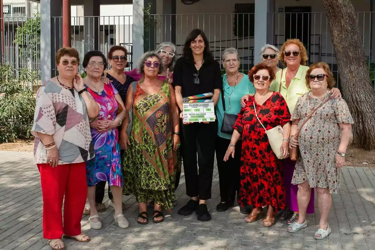 Un grupo de personas posando juntas al aire libre, algunas de ellas llevan gafas de sol y ropa colorida, mientras una persona en el centro sostiene una claqueta del documental Ellas en la ciudad.