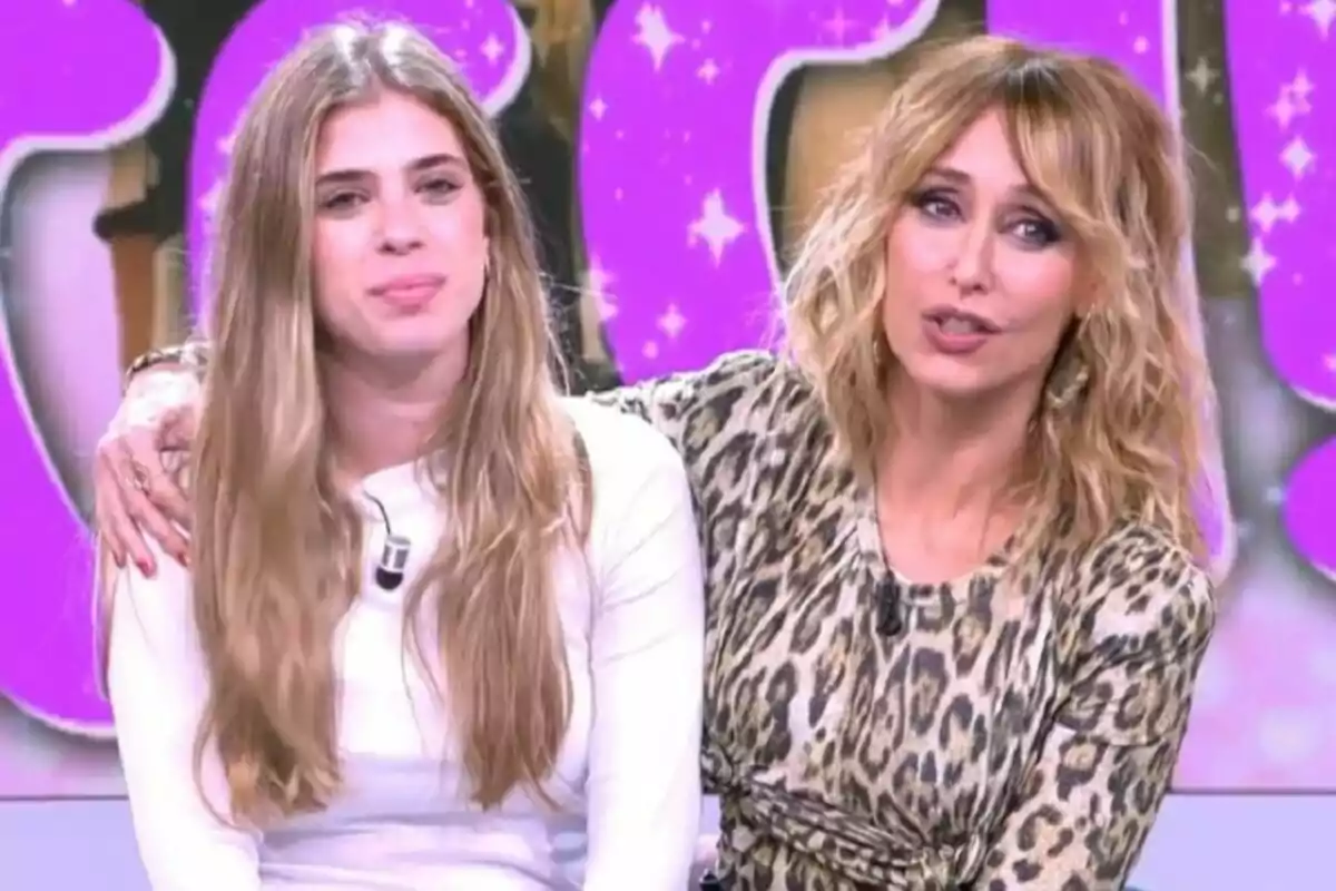 Emma García y Cecilia sentadas juntas en Fiesta, una con cabello largo y rubio y la otra con cabello rizado y un vestido de estampado animal, con un fondo decorado en tonos morados. Emma García y Cecilia sentadas juntas en Fiesta, una con cabello largo y rubio y la otra con cabello rizado y un vestido de estampado animal, con un fondo decorado en tonos morados.