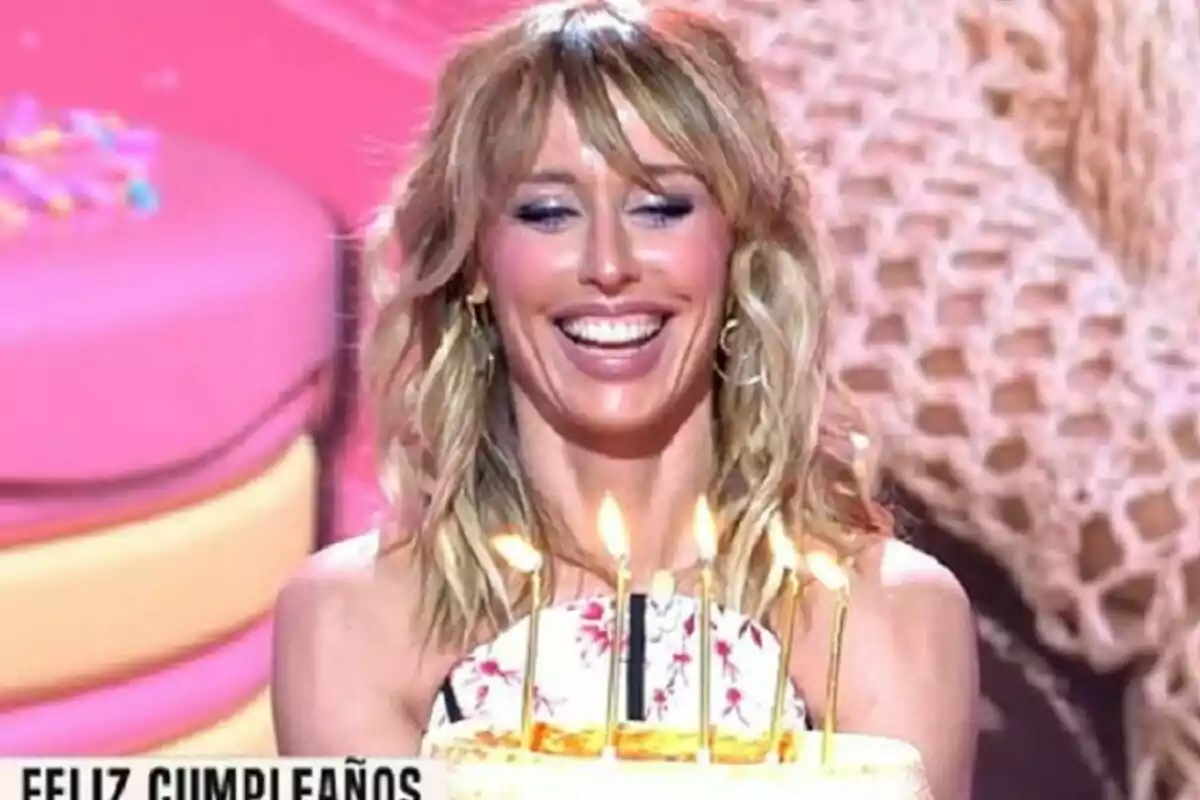 Emma García sonriente sosteniendo un pastel de cumpleaños con velas encendidas frente a un fondo colorido en Fiesta Emma García sonriente sosteniendo un pastel de cumpleaños con velas encendidas frente a un fondo colorido en Fiesta