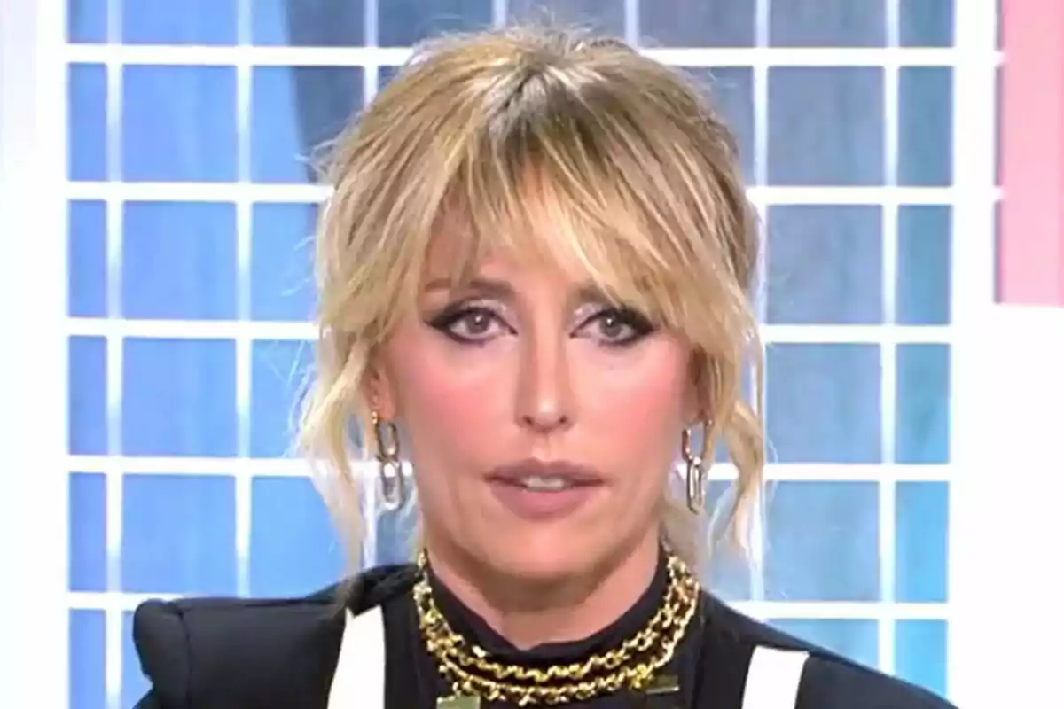 Emma García con cabello rubio y maquillaje oscuro en un set de televisión con fondo de cuadros en Fiesta.