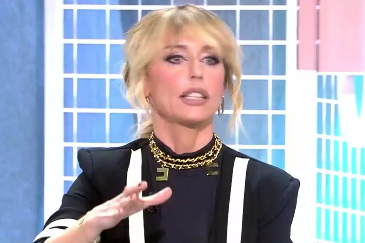 Emma García con cabello rubio y maquillaje pronunciado está hablando en un set de televisión con un fondo de cuadros azules y blancos en Fiesta. Emma García con cabello rubio y maquillaje pronunciado está hablando en un set de televisión con un fondo de cuadros azules y blancos en Fiesta.