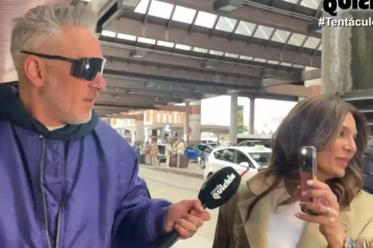 Kiko Hernández con gafas de sol sostiene un micrófono de Tentáculos mientras entrevista a Raquel Bollo que sostiene un teléfono móvil en un entorno urbano con coches y una estructura de techo visible. Kiko Hernández con gafas de sol sostiene un micrófono de Tentáculos mientras entrevista a Raquel Bollo que sostiene un teléfono móvil en un entorno urbano con coches y una estructura de techo visible.