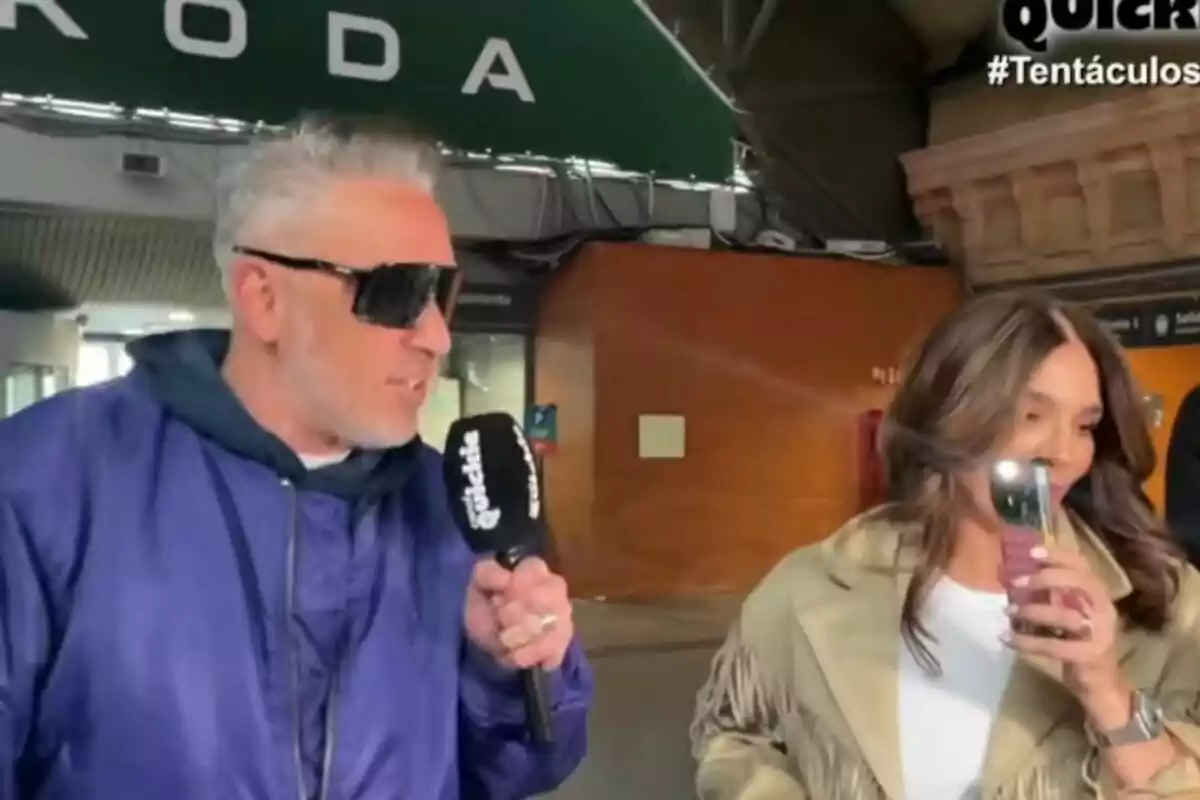 Kiko Hernández con gafas de sol sostiene un micrófono de Tentáculos mientras Raquel Bollo a su lado toma una foto con su teléfono móvil.