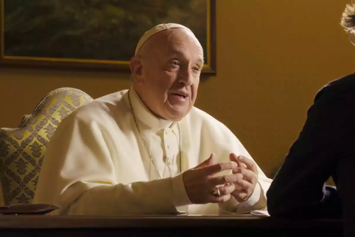 El Papa Francisco vestido con atuendo religioso blanco está sentada en una silla decorada, gesticulando con las manos mientras conversa con alguien fuera de la imagen.