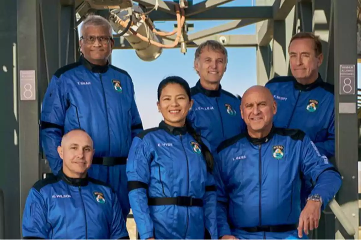 Un grupo de seis personas vestidas con trajes espaciales azules posando juntas en una estructura metálica al aire libre.