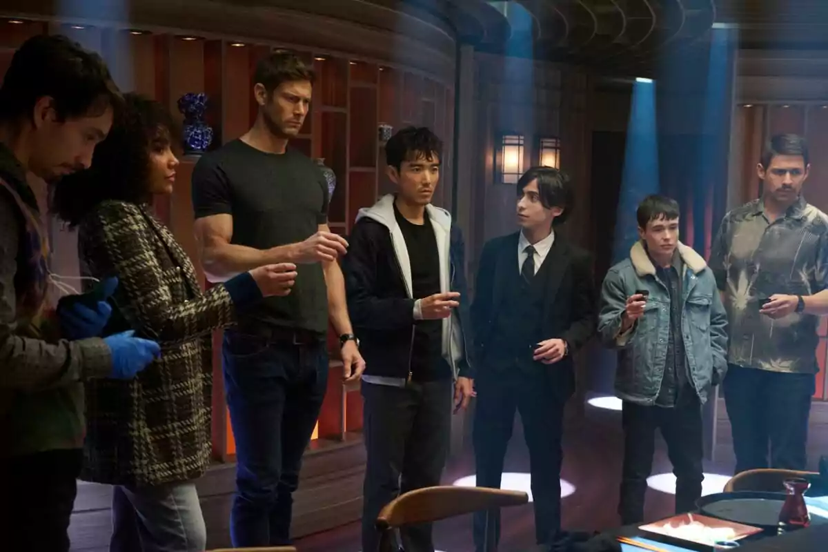 Un grupo de personas de pie en una habitación con iluminación tenue, algunas sosteniendo pequeños objetos en sus manos en una escena de la última temporada de la serie The Umbrella Academy Un grupo de personas de pie en una habitación con iluminación tenue, algunas sosteniendo pequeños objetos en sus manos en una escena de la última temporada de la serie The Umbrella Academy