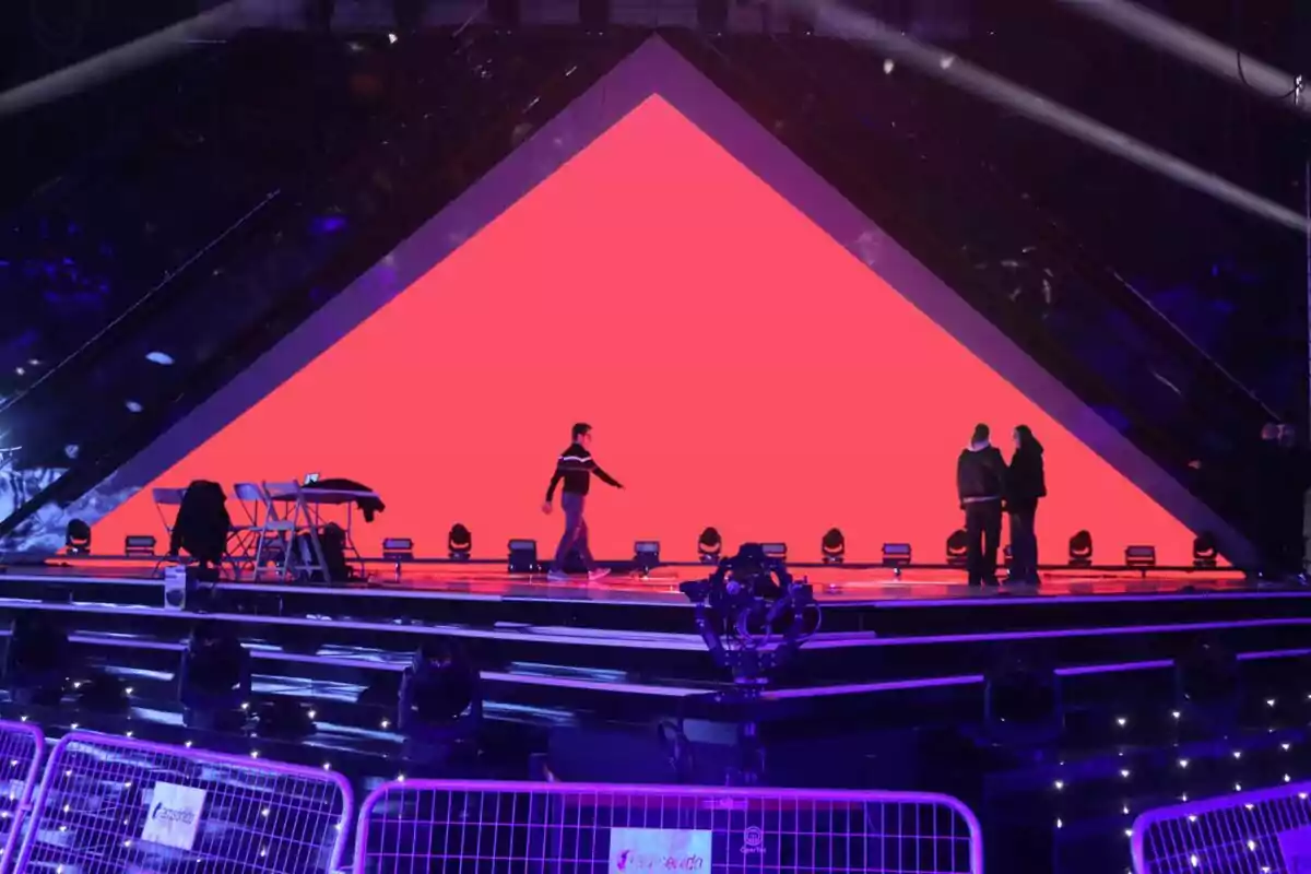Un escenario iluminado del Benidorm Fest con una gran pantalla roja triangular al fondo y varias personas caminando y conversando. Un escenario iluminado del Benidorm Fest con una gran pantalla roja triangular al fondo y varias personas caminando y conversando.