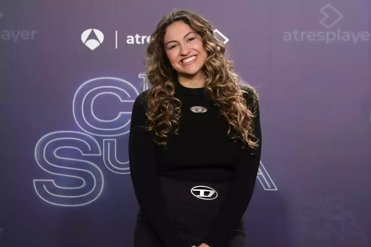 Esperansa Grasia con cabello rizado y suelto sonríe frente a un fondo con logotipos y texto de Tu Cara Me Suena. Esperansa Grasia con cabello rizado y suelto sonríe frente a un fondo con logotipos y texto de Tu Cara Me Suena.