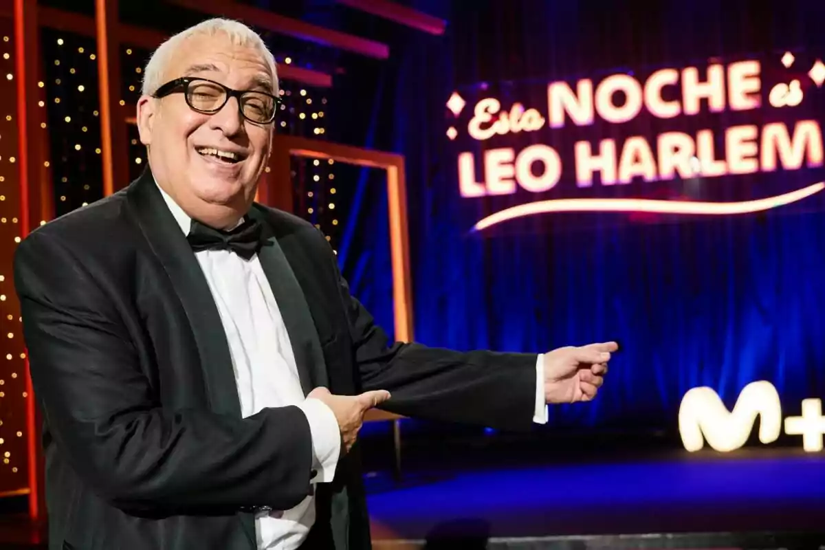 Leo Harlem con gafas y esmoquin sonríe mientras señala un letrero luminoso que pone Leo Harlem con gafas y esmoquin sonríe mientras señala un letrero luminoso que pone