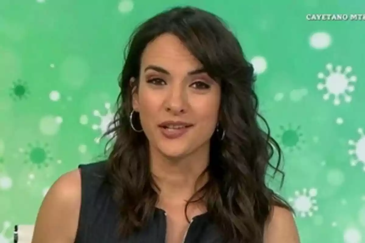 Esther Vaquero con cabello oscuro y rizado usando aretes grandes frente a un fondo verde con patrones circulares en Antena 3 Noticias. Esther Vaquero con cabello oscuro y rizado usando aretes grandes frente a un fondo verde con patrones circulares en Antena 3 Noticias.