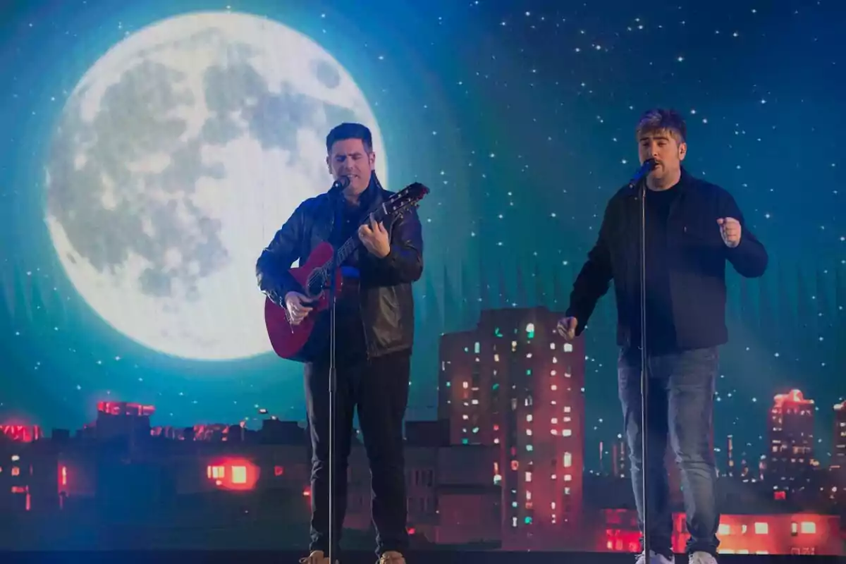 Estopa en un escenario con una guitarra y un micrófono, con un fondo de luna llena y edificios iluminados en el especial de Nochebuena de RTVE. Estopa en un escenario con una guitarra y un micrófono, con un fondo de luna llena y edificios iluminados en el especial de Nochebuena de RTVE.