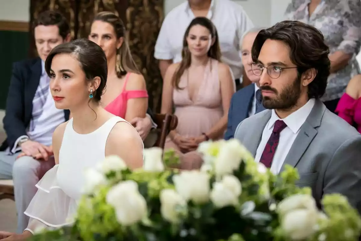 Una pareja elegantemente vestida está sentada en un evento formal rodeada de otras personas, con un arreglo de flores en primer plano en Eternamente amándonos. Una pareja elegantemente vestida está sentada en un evento formal rodeada de otras personas, con un arreglo de flores en primer plano en Eternamente amándonos.