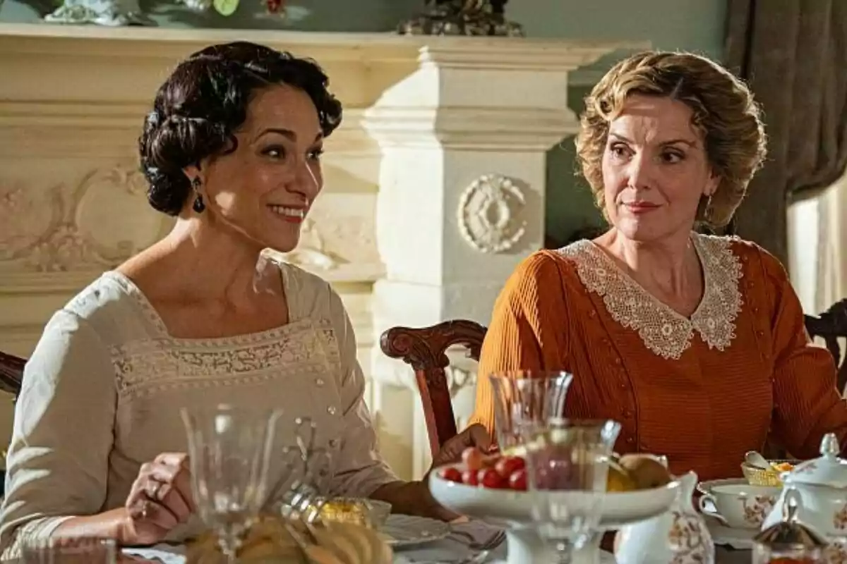 Dues dones assegudes a taula en una escena d'època, que són Eva Martín i Isabel Serrano, una somriu mentre l'altra l'observa amb atenció, envoltades de vaixella i menjar. Dues dones assegudes a taula en una escena d'època, que són Eva Martín i Isabel Serrano, una somriu mentre l'altra l'observa amb atenció, envoltades de vaixella i menjar.