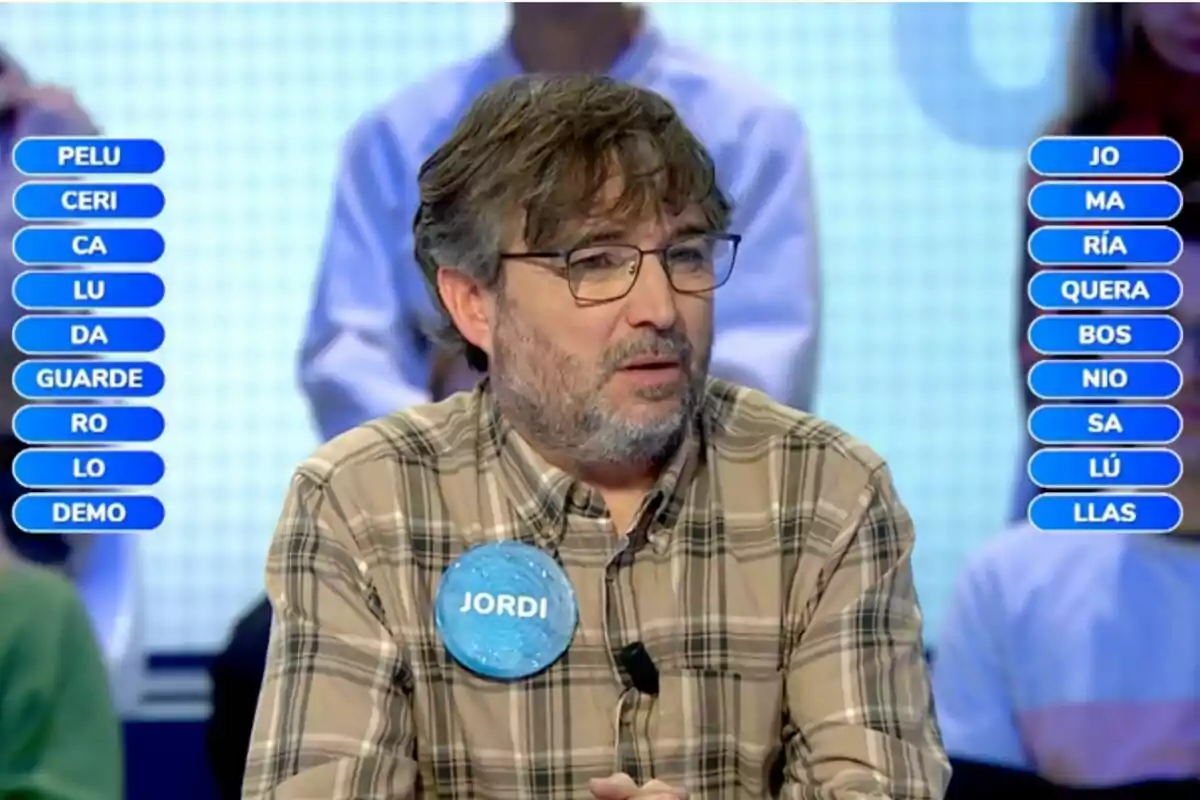 Jordi Évole con gafas y barba, vestido con una camisa de cuadros, participa en 'Pasapalabra'; lleva un cartel con el nombre Jordi Évole con gafas y barba, vestido con una camisa de cuadros, participa en 'Pasapalabra'; lleva un cartel con el nombre