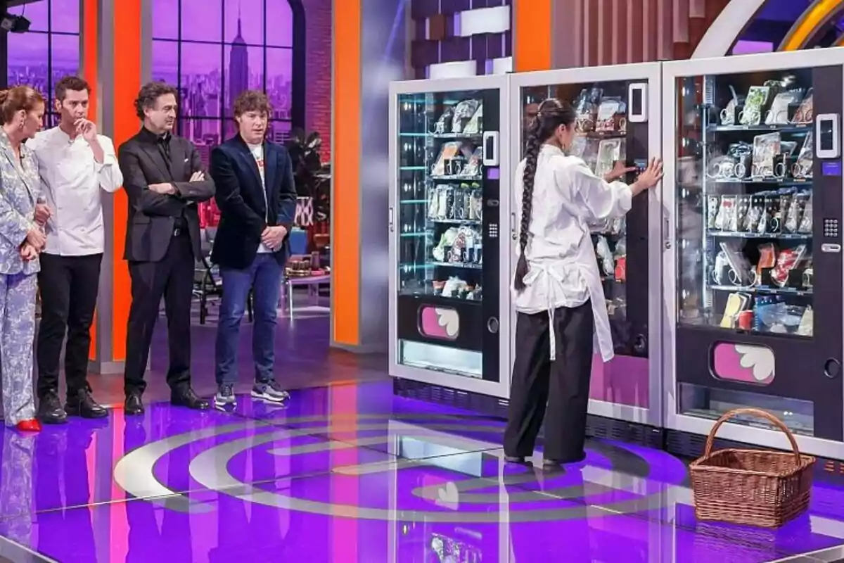 El jurado observa a una mujer que interactúa con una máquina expendedora en un estudio de televisión con un suelo brillante y un fondo colorido en 'Masterchef 12+1'. El jurado observa a una mujer que interactúa con una máquina expendedora en un estudio de televisión con un suelo brillante y un fondo colorido en 'Masterchef 12+1'.