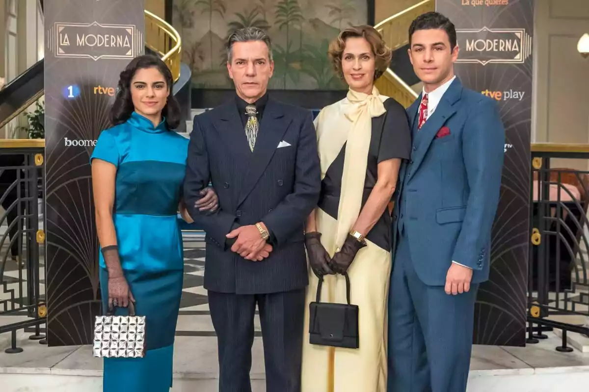 Un grupo de cuatro personas elegantemente vestidas posando frente a un fondo decorativo con el letrero Un grupo de cuatro personas elegantemente vestidas posando frente a un fondo decorativo con el letrero