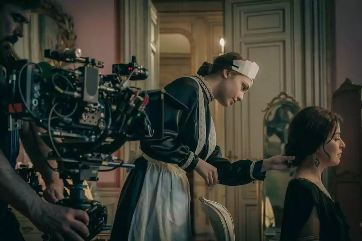 Una escena de rodatge mostra a Verónica Sánchez vestida d'època sent pentinada per una altra persona mentre un càmera grava l'acció en un elegant set.