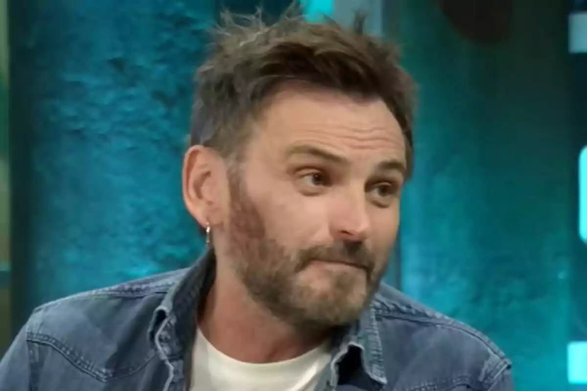 Fernando Tejero con barba y cabello corto lleva una chaqueta de mezclilla y una camiseta blanca mientras está en un entorno con fondo azul en La Revuelta.